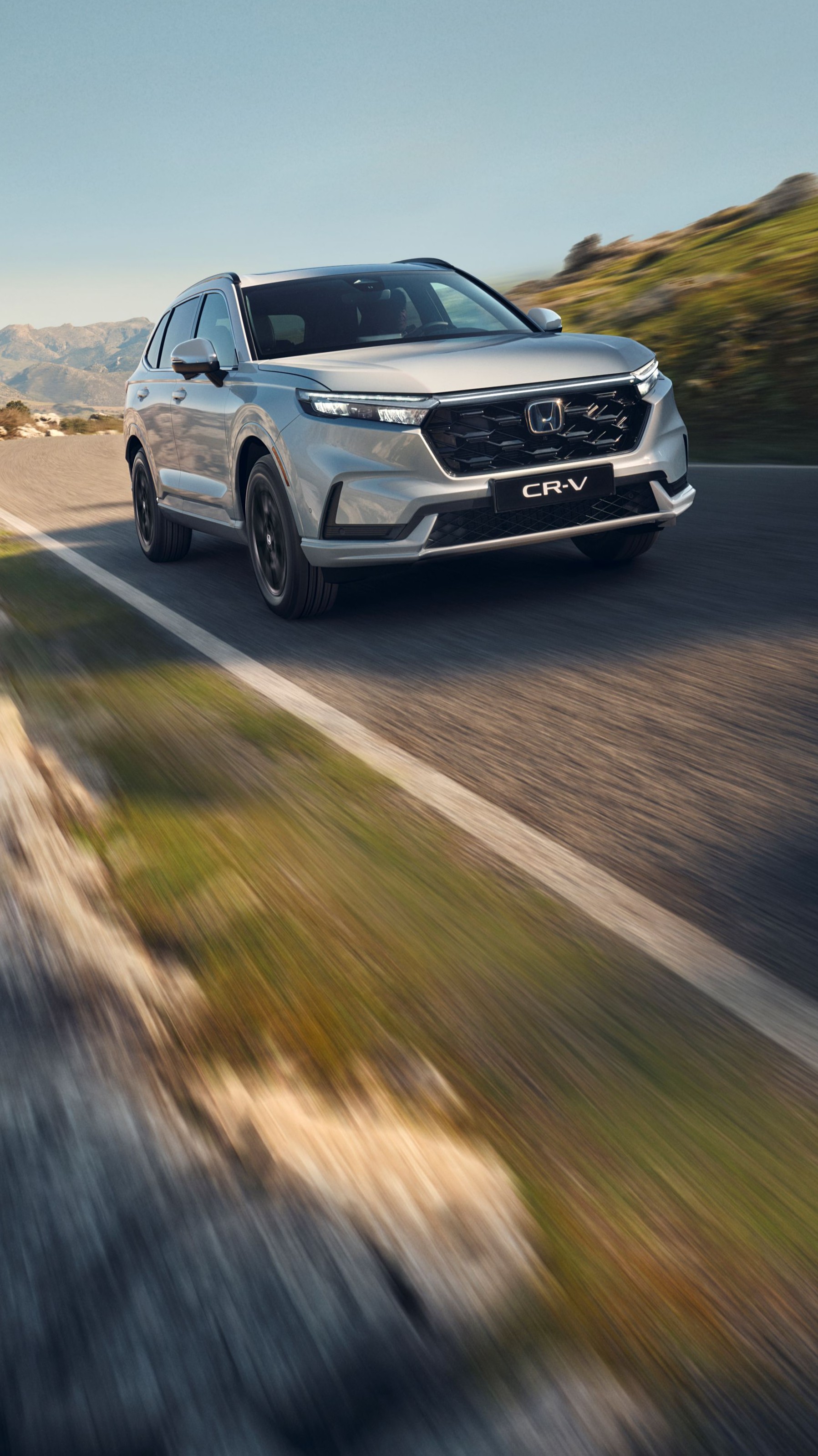 Un SUV Honda CR-V argenté roulant sur une route de montagne sinueuse avec un terrain rocheux et des montagnes en arrière-plan. Le véhicule est en mouvement, créant une sensation de vitesse et d'aventure dans ce paysage escarpé.