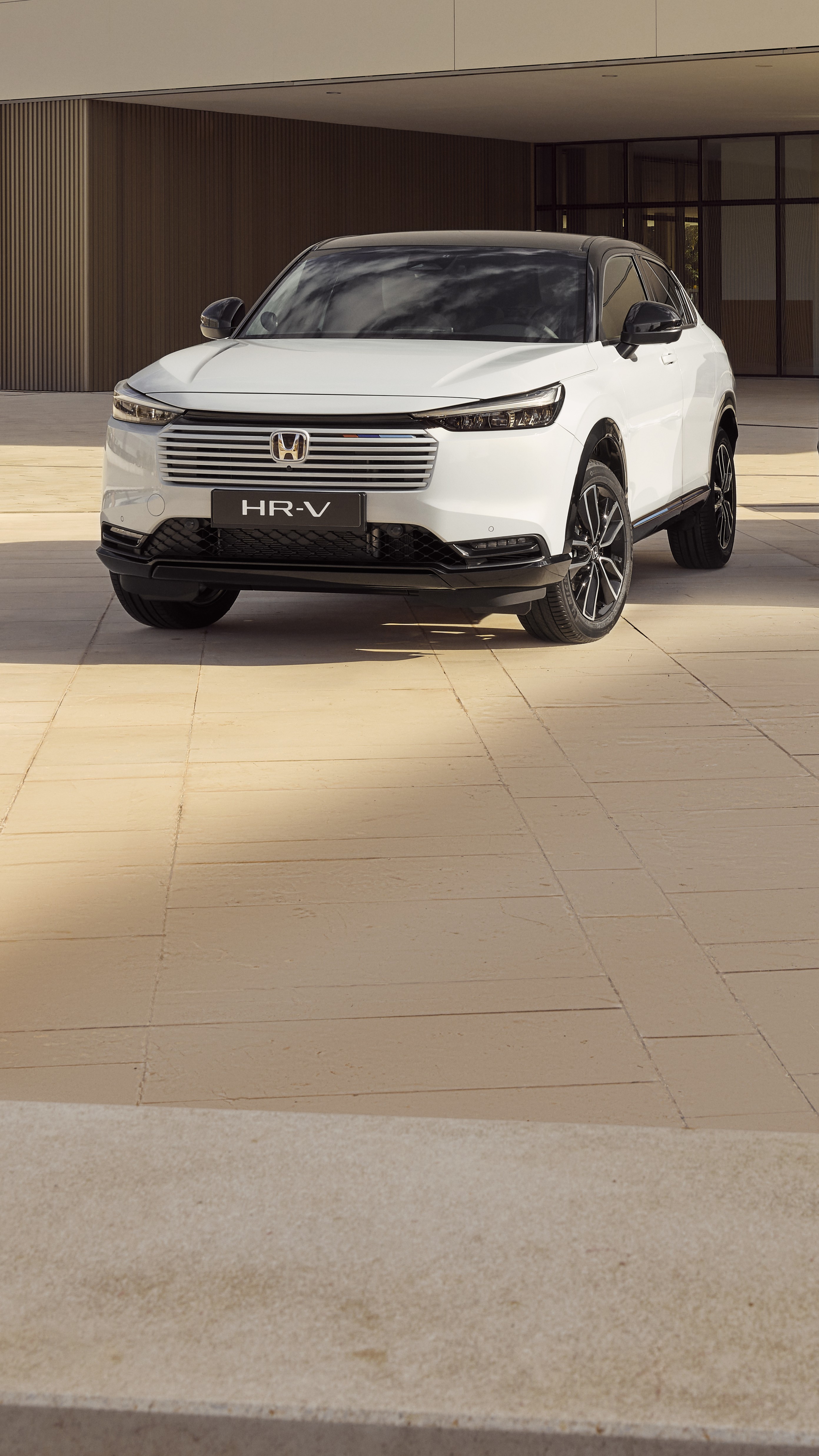 Un SUV Honda HR-V blanc stationné devant un bâtiment moderne aux murs en carrelage beige. Le véhicule est positionné en angle, mettant en valeur son design élégant et sa calandre avant distinctive.