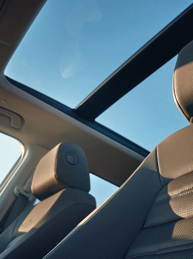 Vue intérieure d'une voiture avec un toit panoramique en verre laissant entrer le ciel bleu et la lumière du soleil, avec des sièges avant en cuir et des appuie-tête illuminés par la lumière naturelle.