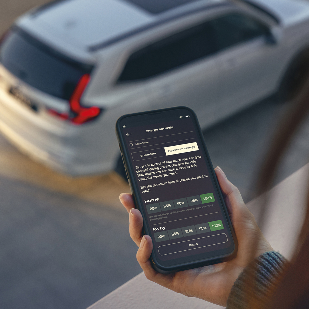 Une personne tient un smartphone affichant une application de configuration de charge pour véhicule, tandis qu'un SUV blanc est garé en arrière-plan.