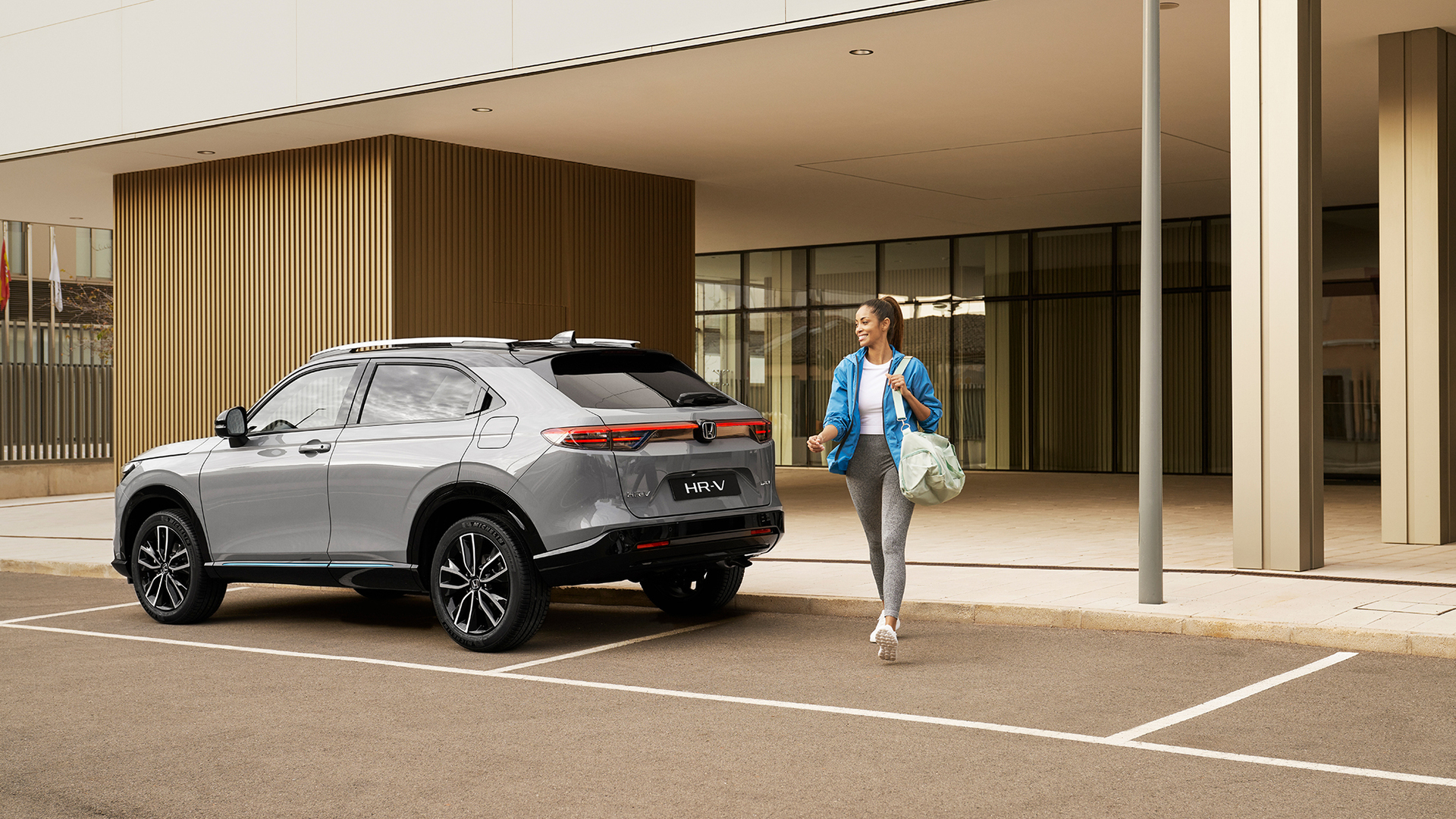 Un Honda HR-V argenté est stationné devant un bâtiment moderne, tandis qu’une femme passe avec un sac, évoquant un usage urbain pratique au quotidien.