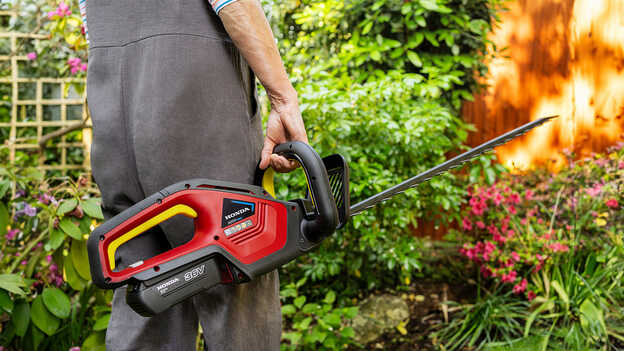 Modèle tenant un taille-haie à batterie Honda dans un jardin.