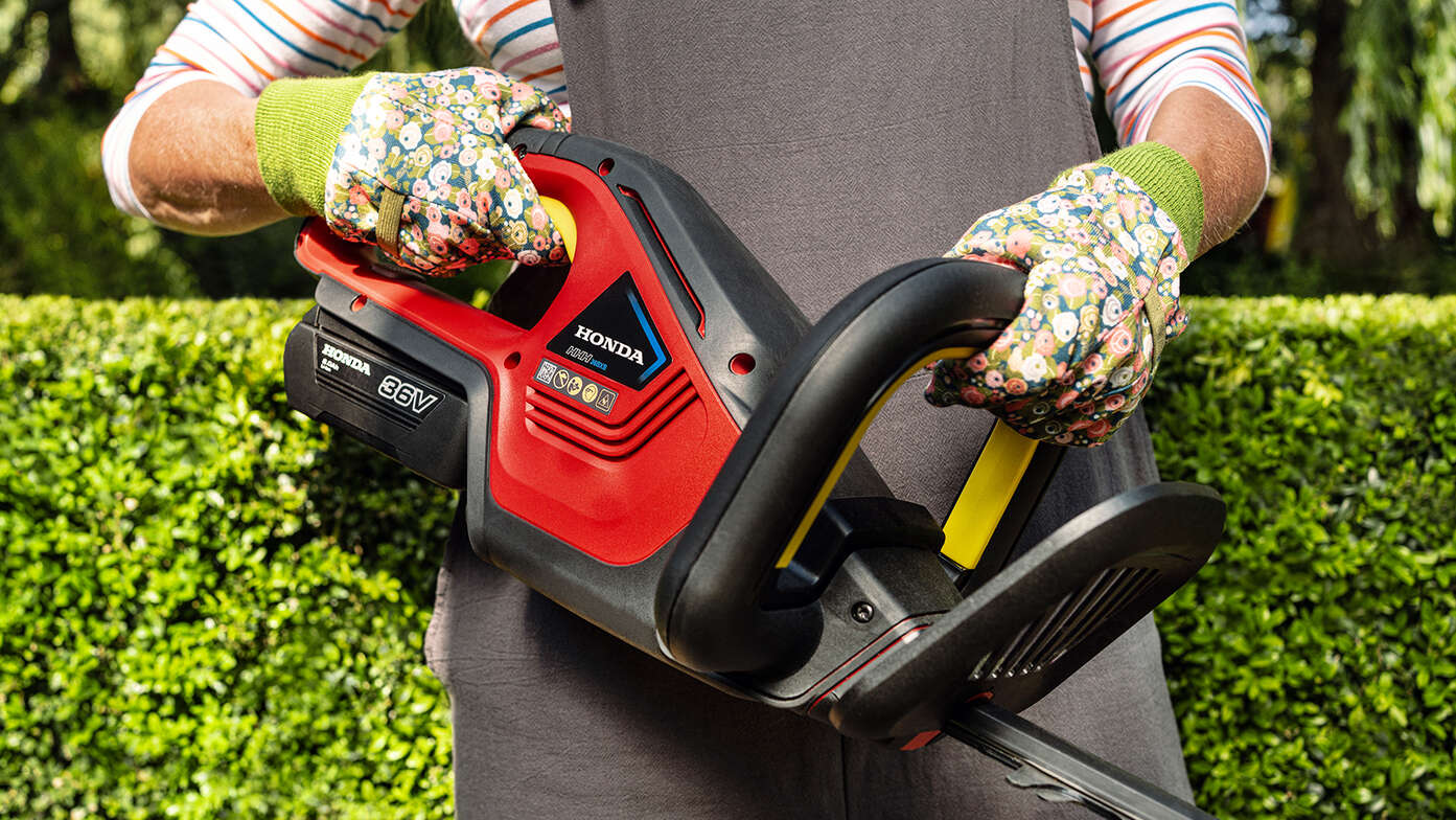 Modèle tenant un taille-haie à batterie Honda par la poignée dans un jardin.