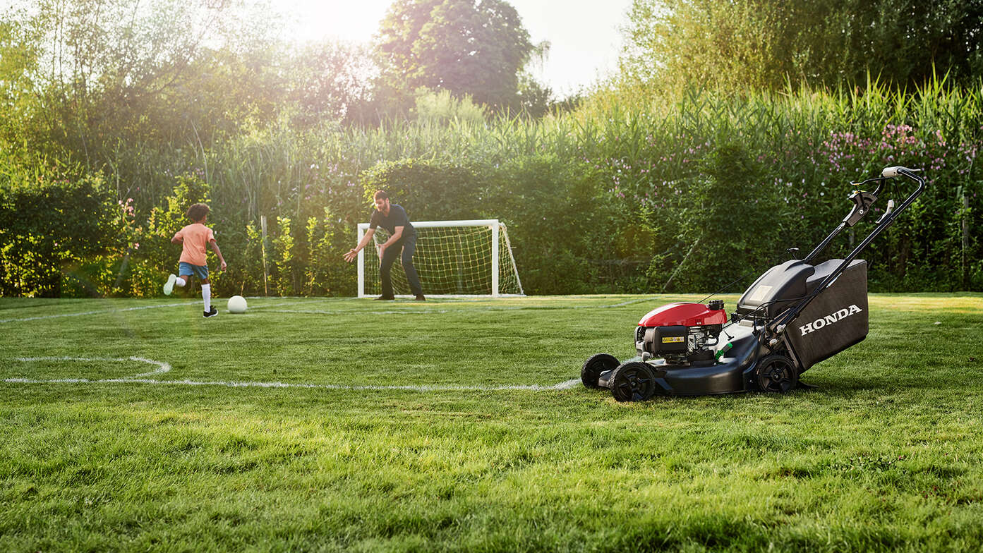 Un homme jouant au football avec un enfant à côté d'une Honda HRN dans le jardin. 