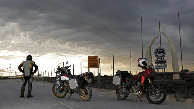 Pilote fixant l'horizon à côté de deux Africa Twin stationnés sur l'asphalte.