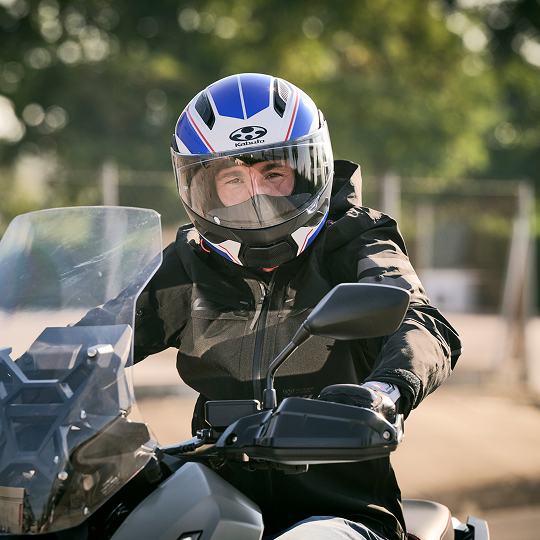 A motorcyclist wearing a blue and white helmet and black jacket sits on a Honda motorcycle with a windshield, ready to ride.
