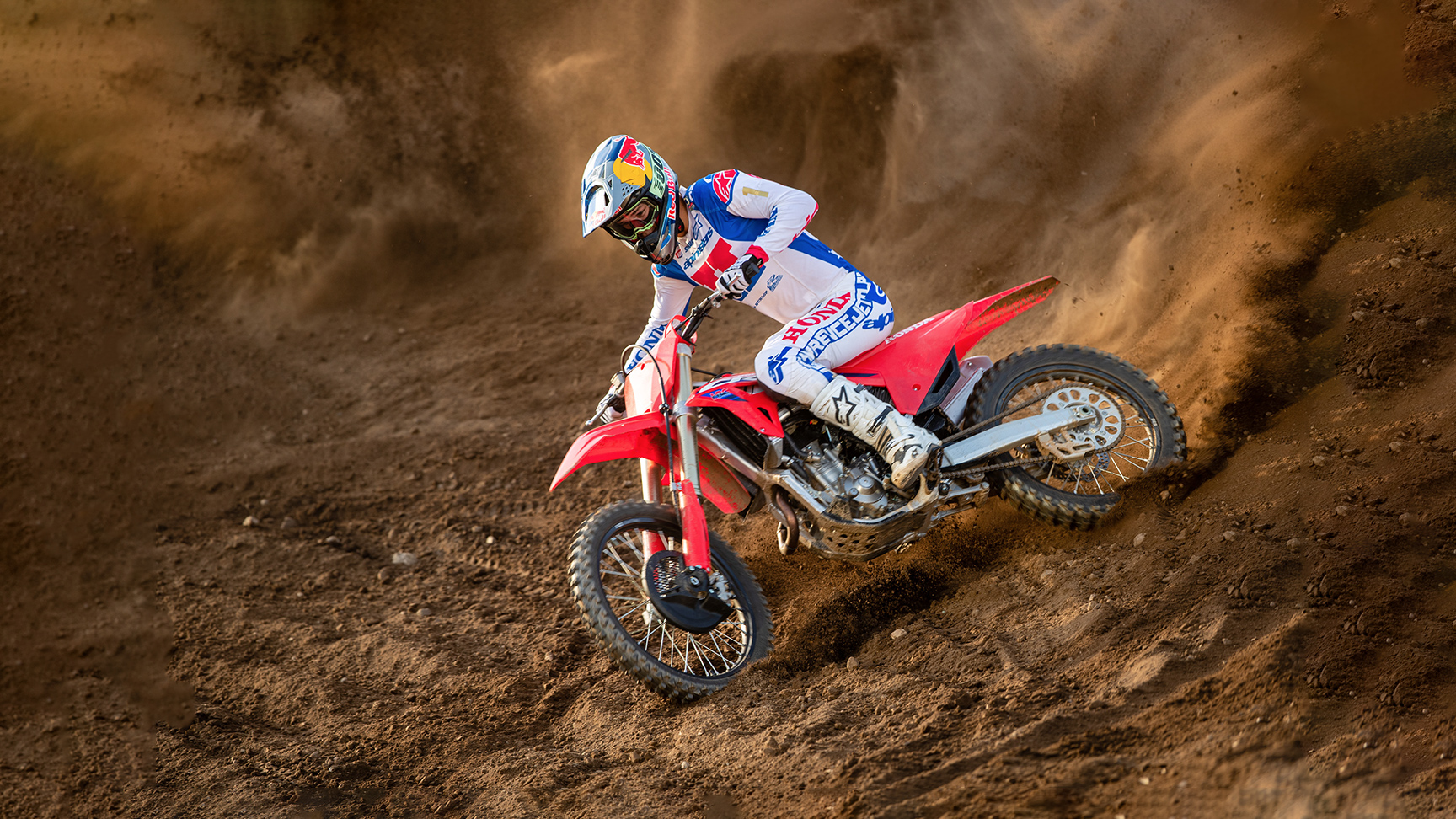 Un pilote de motocross en tenue de course blanche, bleue et rouge prend un virage serré sur une moto tout-terrain rouge, creusant la piste sablonneuse et soulevant un spectaculaire nuage de poussière derrière lui.
