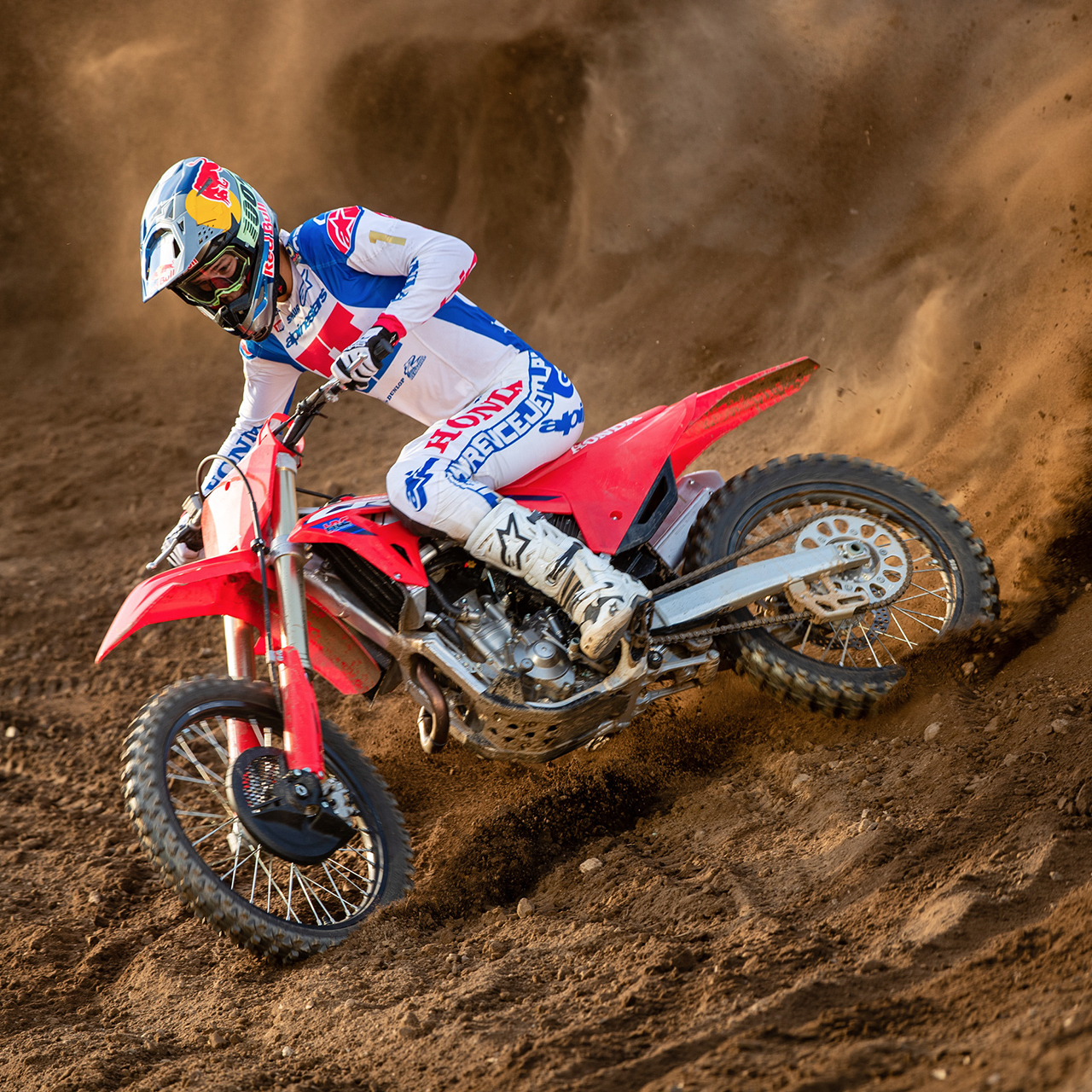 Un pilote de motocross en tenue de course blanche, bleue et rouge prend un virage serré sur une moto tout-terrain rouge, creusant la piste sablonneuse et soulevant un spectaculaire nuage de poussière derrière lui.