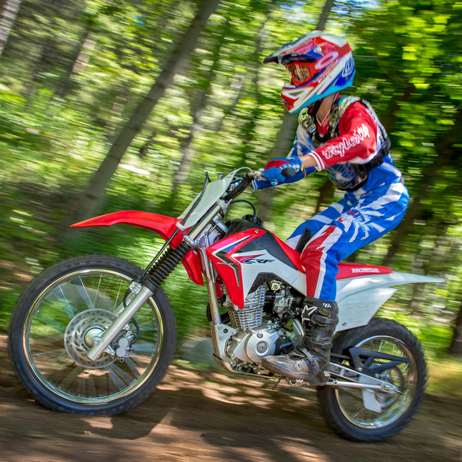 Vue latérale d'une CRF125F avec un pilote en situation sur une piste de forêt. Avant gauche.
