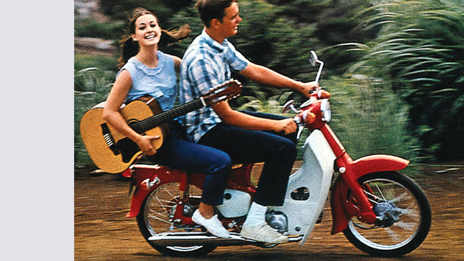 Honda Cub avec conducteur et passager portant une guitare. Chemin en terre. Vue latérale côté droit.