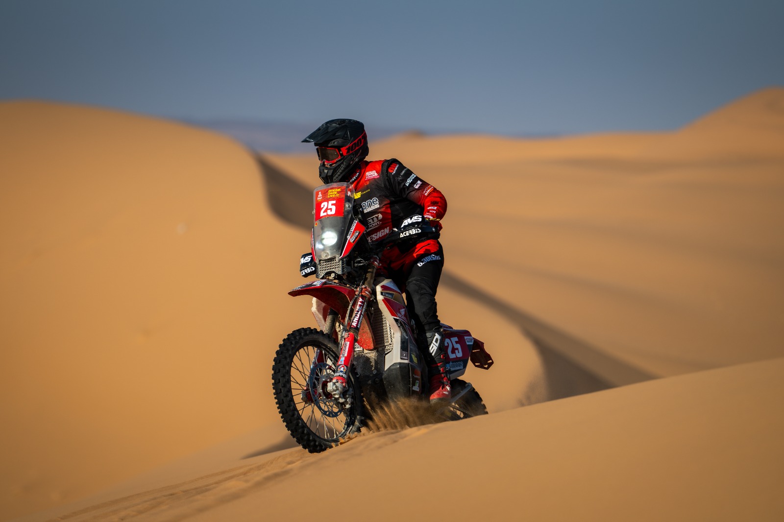A Honda off-road motorcycle rider in red gear navigates sand dunes in a desert landscape. The bike's headlight is on, and the rider is leaning forward, suggesting movement across the challenging terrain.